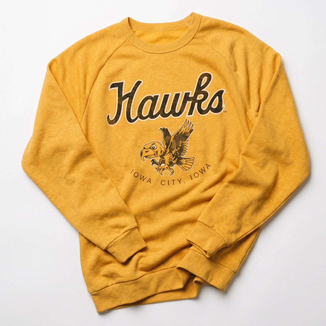 A mustard yellow sweatshirt featuring a dark brown graphic print of the word 'Hawks' in script, an eagle illustration, and 'IOWA CITY, IOWA' underneath, laid flat on a light gray background.